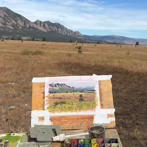 A watercolor painting of a grassy field with distant mountains under a partly cloudy sky, set up outdoors on an easel with paint supplies visible.