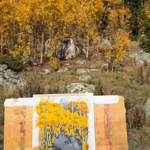 A watercolor painting of golden autumn trees and rocky terrain set against a real autumn forest backdrop, captured during an outdoor painting session.